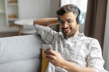 Indian man listening music on smartphone with wireless headphones, relaxing on couch and surfing internet online at home