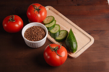 there are three red tomatoes on the table, there is a sliced cucumber on it and a white cup with buckwheat