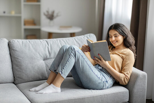 Young Indian woman lying on sofa with open book, enjoying peaceful morning, reading captivating story in living room, copy space