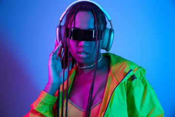 young african american woman in headphones and stylish sunglasses posing in studio with neon lights