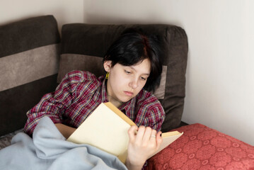 Young teenage girl is concentrating on reading a book lying on sofa at home, enriching her language and learning new foreign language through reading books, concept of self development