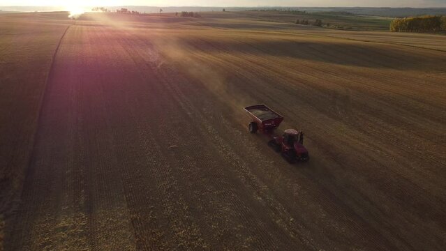Cremona Alberta Canada, October 01 2023: Tractor pulling grain cart across harvested field aerial shot with dust trail at sunset 
