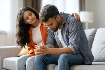 Apologize at home. Indian woman calms offended guy, support and caring, sitting together on sofa in...