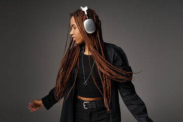 relaxed and young African American woman in 20s with headphones dancing to music on grey backdrop