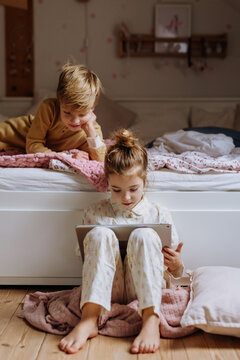 Siblings watching something on tablet, boy lying on bed, cute girl sitting on on floor in bedroom. Children's screen time.