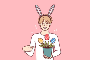 Teenage guy celebrating easter holds pot of plants in shape of multi-colored eggs, wears bunny ears on head. Boy invites to celebrate easter together, or buy decorations to create festive atmosphere