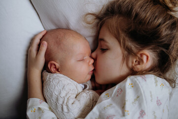 Portrait of big sister cuddling newborn, little baby. Girl lying with her new sibling in bed, kissing baby's cheek. Sisterly love, joy for new family member. - obrazy, fototapety, plakaty