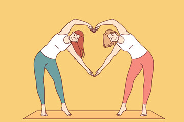 Paired yoga of two women doing aerobics or pilates standing on sports mat and making heart sign from hands. Attractive girls are interested in fitness and pilates to avoid excess weight