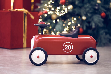 View of red children's retro car on the background of gifts in red boxes with golden bows, Christmas tree decorated garland, string light, red and blue balls. Background, copy space, bokeh.