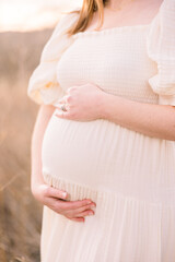 A pregnant mother stands cradling her bump in a field outside at sunset.