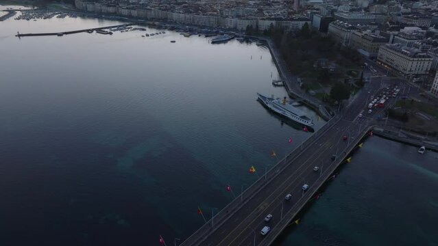 High angle view of cars passing on bridge over water. Tilt up reveal buildings on lake waterfront and sunrise sky. Geneva, Switzerland