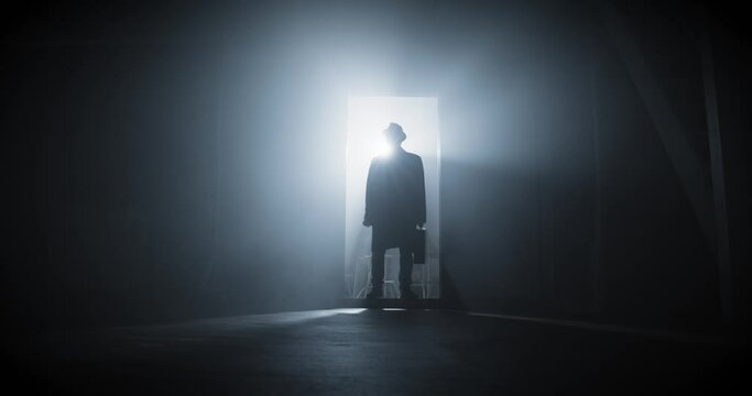 Silhouette of a man dressed in long coat with hat walking to a dark room through a door. Cinematic shot. High quality 4k footage