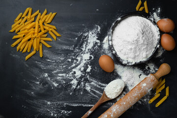 Raw homemade pasta penne on dark background with eggs and flour. Top view.