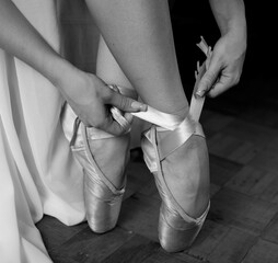 A ballerina in a white dress and pointe shoes sits and puts on pointe shoes on a black background