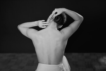 A ballerina in a white dress and pointe shoes sits on the floor with her back to the camera on a black background