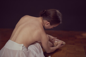 A ballerina in a white dress and pointe shoes sits on the floor with her back to the camera on a black background