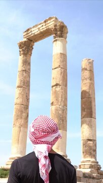 Rear view portrait of young guy with typical head cover in Jordan standing looking at ruins of the Temple of Hercules on the top of the mountain of the Amman citadel.