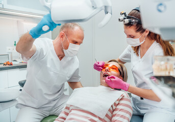 Dental clinic patient appointment. Dentist woman in magnifying glasses doing teeth prevention using medical tools. Young man assistant pointing light. Health care and medicare industry concept image.