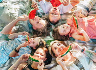 Young daughters with parents family lying on picnic blanket during weekend sunny day, smiling, laughing and eating red juicy watermelon pieces. Family values, fruits vitamins, outdoor time concept