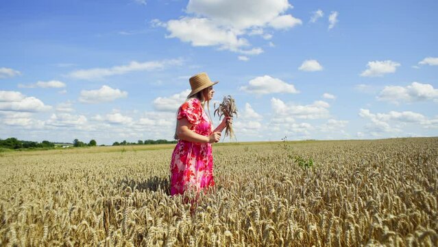 A woman in a beautiful dress with a bouquet of ears of corn in her hands dances. She enjoys her life in the middle of a large wheat field. High quality 4k footage