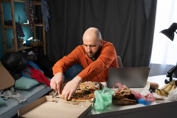 Millennial man eating testy pizza while working or studying in messy room
