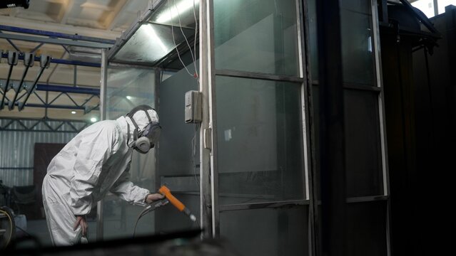 A worker in protective overalls works with hazardous chemicals, spraying them onto metal. Close-up of a worker wearing a respirator in a paint shop. 