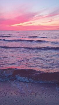 Vertical footage video 4K of evening relax on beach. Calming motion of small sea waves break on sand. Sunset pink sky reflects in water at golden hour. Meditative walk on ocean coast. Peaceful mood