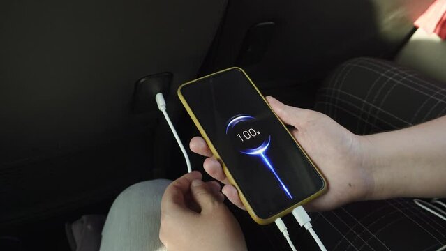 Woman connects charging cable of smartphone to USB socket in seat of tourist bus. Phone shows full charge while traveling. Interior of public comfortable service for international transport.