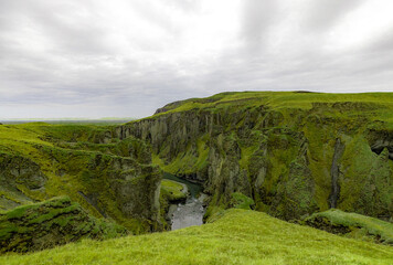 A river meanders through a canyon surrounded by two towering mountains, with a clear blue sky overhead and lush green grass covering the slopes. Fjadrargljufur Canyon