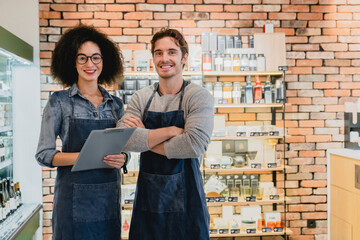 Two smiling helpful shop assistants standing close to goods shelves holding folder against brick wall. Multi-cultural sellers shop owners cafeteria staff waiters posing for camera