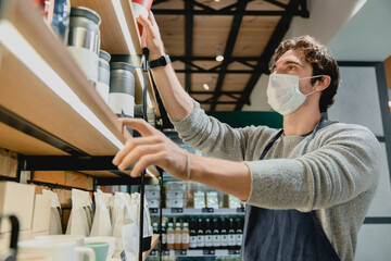 Young attractive waiter in medical mask against coronavirus taking glasses from the shelf in coffee shop. Barista bartender brewing coffee to customers clients in cafeteria