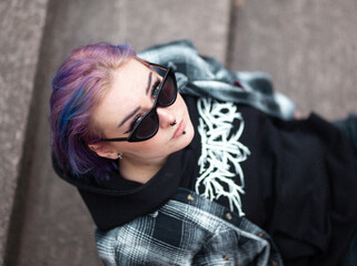 Young woman with colorfull blue hair, piercings and sunglasses posing. Wearing flannel and hoodie. Urban background.