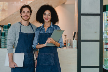 Confident young multicultural cafe workers holding notepads in coffee shop. Shop assistants...