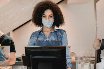 Close up portrait of young helpful african waitress in medical mask against coronavirus standing at the cafe counter in coffee shop. Pandemic and reopening of small business, selling food and drinks