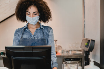 Young african waitress wearing medical mask to protect from covid 19 working at cash point in cafe....
