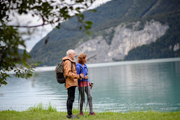 Portrait of beautiful active elderly couple hiking together in autumn mountains. Senior tourists looking at lake. - obrazy, fototapety, plakaty