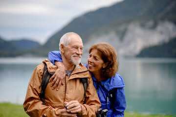 Portrait of beautiful active elderly couple hiking together in autumn mountains. Senior tourists embracing each other in front of lake. - obrazy, fototapety, plakaty