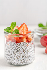 Chia pudding sweet dessert made with seeds soaked in plant based milk with sliced strawberry topping and mint leaf served in glass jar on  on white wooden table with ingredients for healthy breakfast