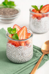 Sweet vegan chia pudding dessert made with seeds soaked in plant based milk with sliced juicy strawberry topping and mint leaf served in glass jar on green napkin on white wooden table with spoon 
