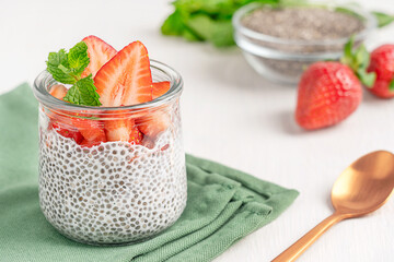 Homemade vegetarian chia pudding made with seeds soaked in plant based milk with sliced strawberry topping and mint leaf served in glass jar on green napkin on white wooden table with spoon as dessert
