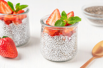 Healthy vegetarian chia pudding made with seeds soaked in plant based milk decorated with sliced strawberry topping and fresh mint leaf served in glass jar on white wooden table with spoon for lunch
