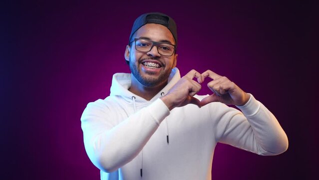 A joyful biracial man in a white hoodie and cap forms a heart with his hands, sharing a universal gesture of love and happiness on a purple backdrop. Camera 8K RAW. 