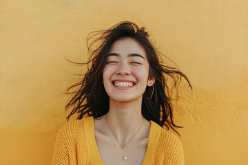 Joyful young woman smiling with hair blowing in the wind, against a yellow textured wall.