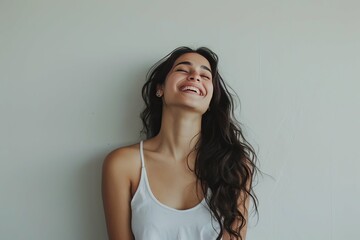 Joyful young woman smiling with closed eyes, leaning against a light grey background.