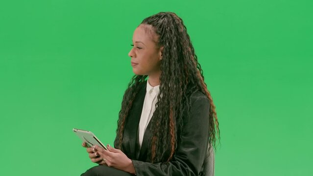 Young female reporter isolated on chroma key green screen background. African american woman tv news host sit in chair with tablet taking interview.