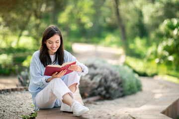 Contented young woman deeply engrossed in reading a red book, sitting with legs crossed on a park...