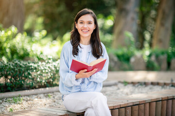 Enthusiastic young reader sits on a park bench with a beaming smile, engrossed in a captivating red...