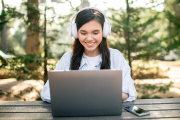 A young woman with headphones smiles while using a laptop at an outdoor wooden table