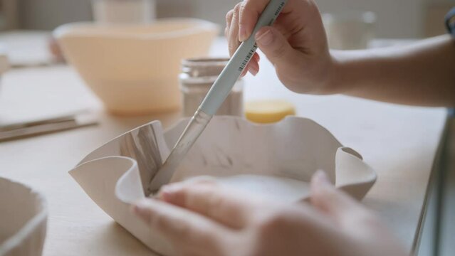 young Asian artisan sits at table in ceramic studio. female ceramist working at craft table desk, glazing and painting clayware, using brush and can. young art woman crafting using clay tools