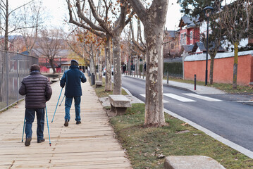 Two men are walking down a sidewalk with the walking sticks or trekking poles. The sidewalk is lined with trees and there is a bench on the sidewalk. The street is empty and there are no other people 